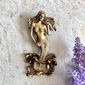 May include: A gold-toned mermaid figurine with a flowing tail and a seashell bra. The mermaid is standing on a wave-like base with coral details.