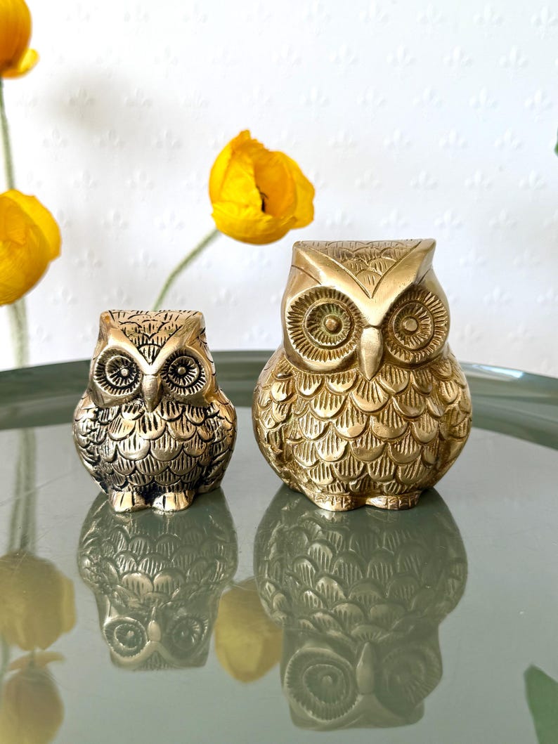May include: Two gold metal owl figurines. The larger owl is about 3 inches tall and the smaller owl is about 2 inches tall. Both owls have detailed features and are made of a shiny metal.