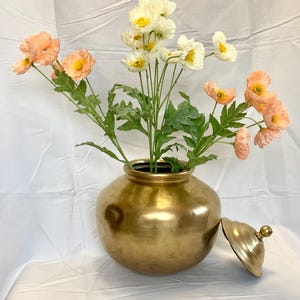 May include: A gold-colored metal vase with a lid, filled with artificial flowers. The flowers are a mix of white and peach-colored poppies with green stems and leaves. The vase is round with a wide base.