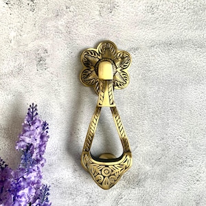 Pode incluir: Um batente de porta dourado com um design floral ornamentado. O batente tem a forma de uma lágrima com um anel na parte superior.