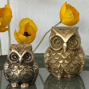 May include: Two gold-colored owl figurines with detailed feather patterns. The larger owl is on the right and the smaller owl is on the left.