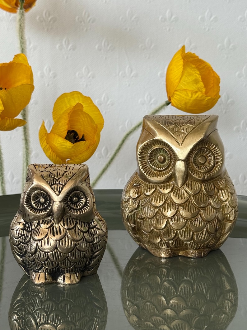 May include: Two gold-colored owl figurines with detailed feather patterns. The larger owl is on the right and the smaller owl is on the left.
