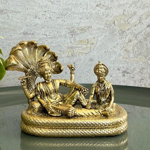 Op de afbeelding: Een goudkleurig beeld van een liggende figuur met meerdere armen, versierd met sieraden en een slangenkap. Een kleiner figuur zit in de buurt. Het beeld staat op een basis met een slangachtig ontwerp.