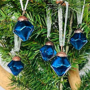 May include: Five blue glass ornaments with a diamond shape and copper tops hanging on a green Christmas tree with white ribbon.