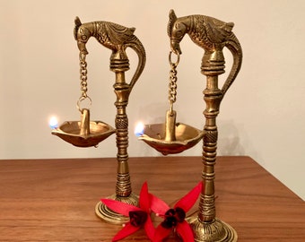 May include: Two brass candle holders with bird-shaped tops and small bowls for oil. The candle holders are lit, with flames visible. Red flowers are placed in front of the candle holders. The candle holders are on a wooden surface.
