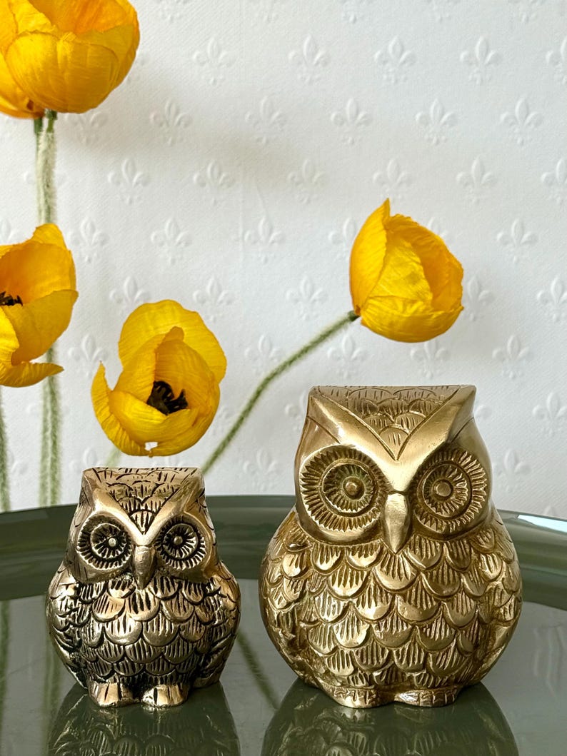 May include: Two gold metal owl figurines. The larger owl is sitting on a green surface. The smaller owl is sitting in front of the larger owl.