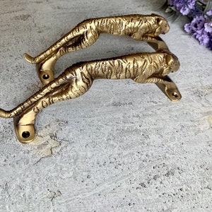 May include: A pair of gold-toned metal door handles shaped like tigers. The handles are detailed with tiger stripes and have a realistic look.
