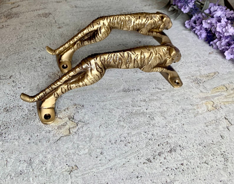 May include: A pair of gold-toned metal door handles shaped like tigers. The handles are detailed with tiger stripes and have a realistic look.