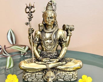 May include: A gold-colored statue of a seated deity, holding a trident and a drum. The statue is adorned with intricate jewelry and a serpent. Two yellow flowers are placed near the statue.