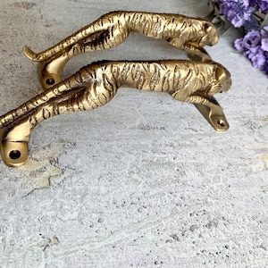 May include: Two gold-toned metal door handles shaped like running tigers. The handles are mounted on a white surface.