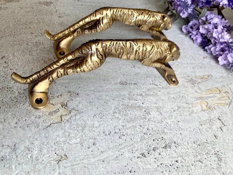 May include: Two gold-toned metal door handles shaped like running tigers. The handles are mounted on a white surface.