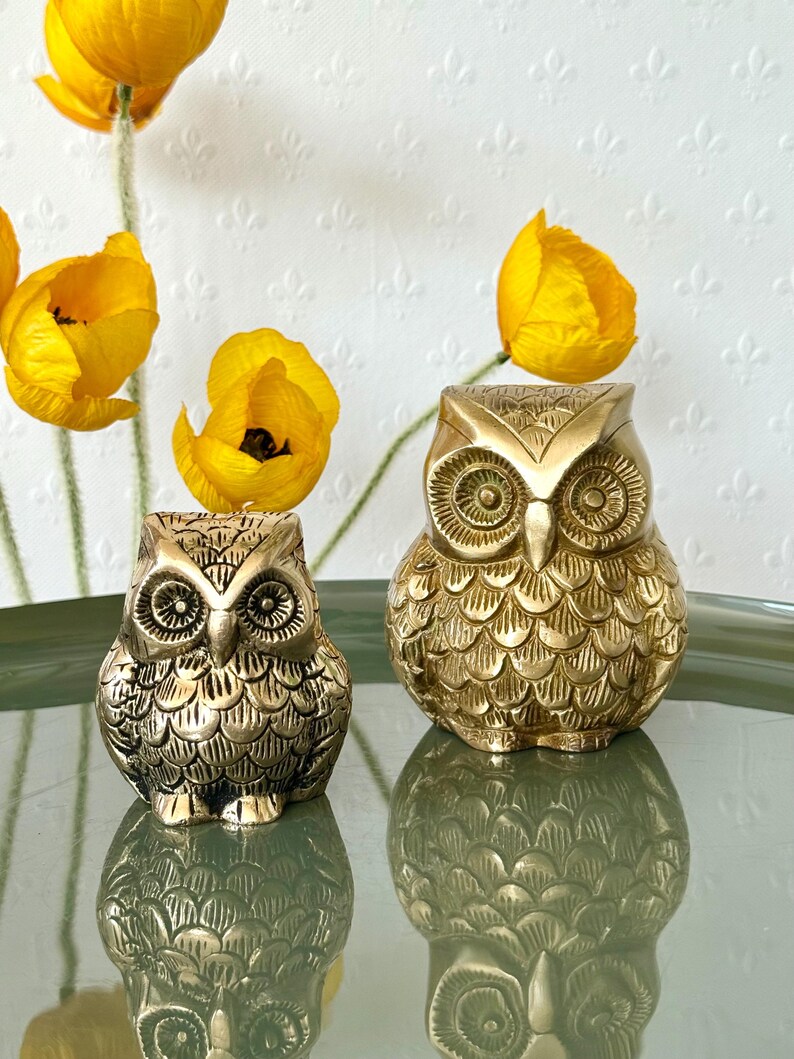 May include: Two gold owl figurines with detailed feather patterns. The larger owl is slightly taller than the smaller owl.