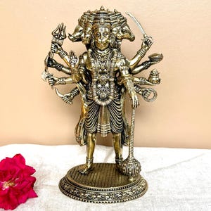 May include: A detailed bronze-coloured statue of a deity with multiple arms and heads, holding various objects. The statue stands on a decorative base and features intricate detailing. A red rose is visible in the foreground.