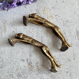 May include: Two gold-toned metal tiger doorstops. The tigers are in a leaping pose with their heads turned to the side. The doorstops are detailed with stripes and have a flat base.