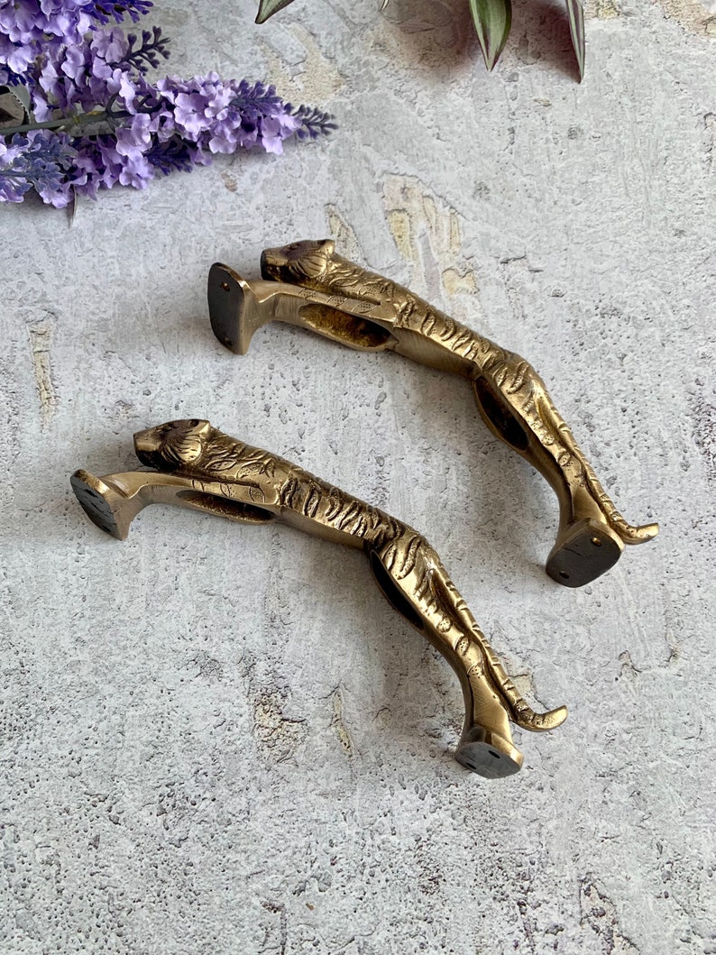 May include: Two gold-toned metal tiger doorstops. The tigers are in a leaping pose with their heads turned to the side. The doorstops are detailed with stripes and have a flat base.