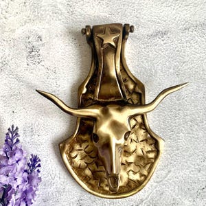 May include: A gold-toned brass door knocker shaped like a bull's head with a star on the top. The knocker has a textured surface and a detailed design.