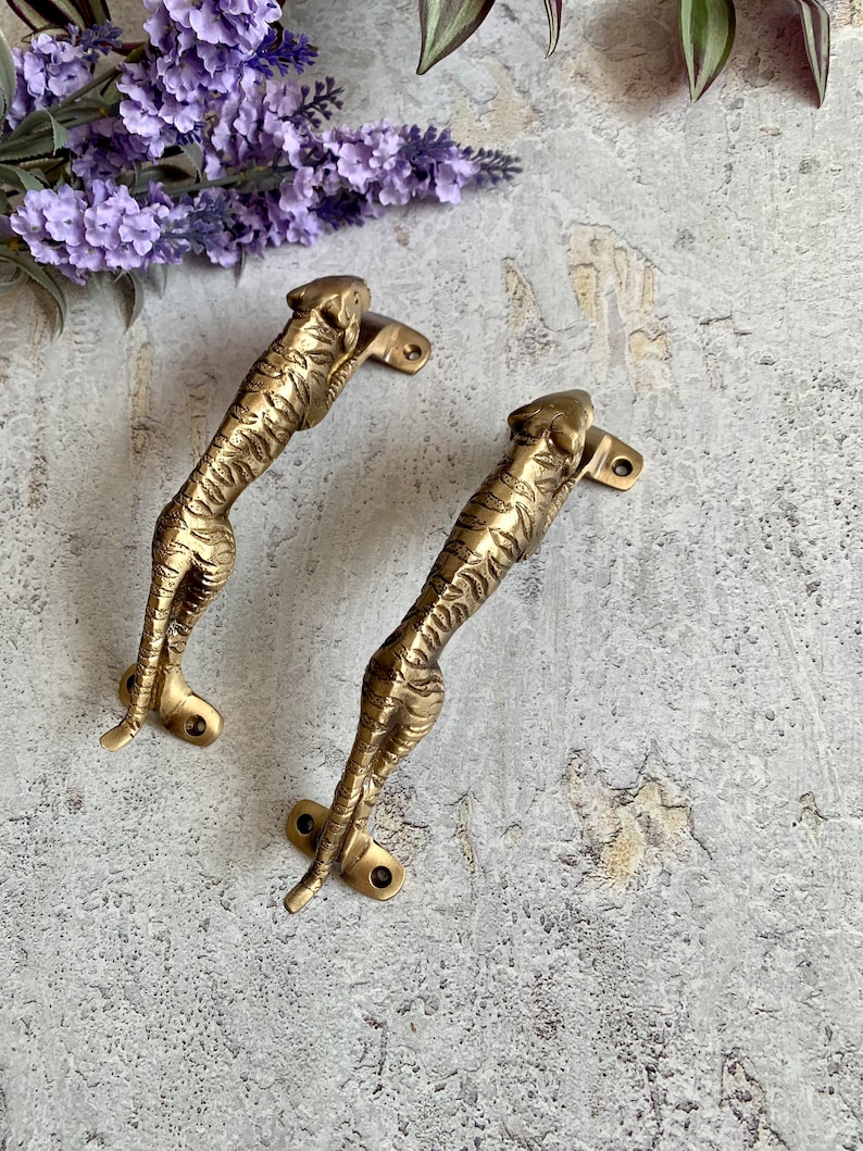 May include: Two gold-toned tiger-shaped door handles with detailed features. The handles are mounted on a white surface.