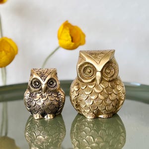 May include: Two gold owl figurines, one larger than the other, with detailed feather patterns and large, round eyes. The owls are sitting on a green surface.