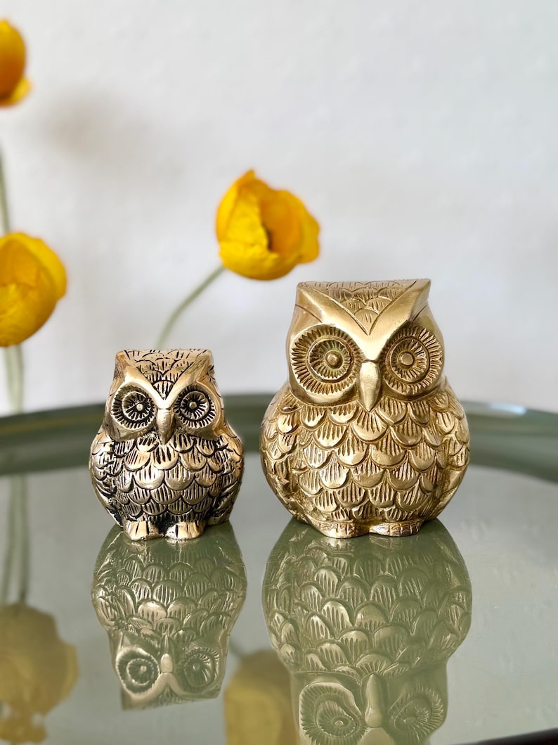 May include: Two gold owl figurines, one larger than the other, with detailed feather patterns and large, round eyes. The owls are sitting on a green surface.