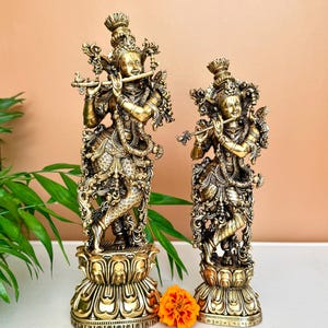 May include: Two ornate brass statues of figures playing flutes, each standing on a detailed lotus-shaped base. The figures are adorned with intricate jewelry, elaborate headpieces, and flowing garments, captured in a metallic gold finish.