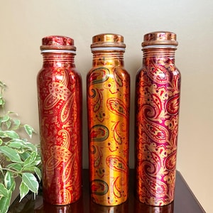 May include: Three copper water bottles with a paisley design in red, gold, and maroon. The bottles have a cylindrical shape with a screw-on cap. The bottles are approximately 25 cm tall.