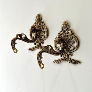 Puede incluir: Dos ganchos de pared de metal dorado adornados con un diseño de volutas.