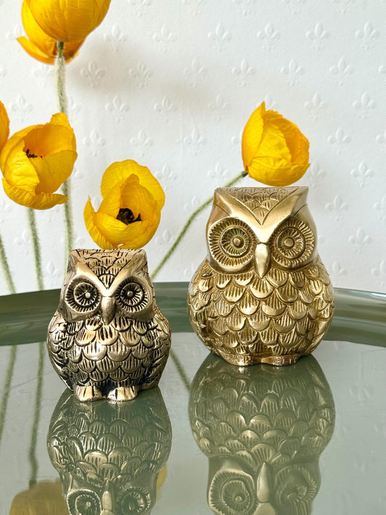 May include: Two gold owl figurines with detailed feather patterns. The larger owl is slightly taller than the smaller owl.
