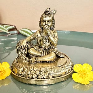 Puede incluir: Una estatua dorada de una figura sentada, adornada con detalles intrincados, incluyendo una corona y joyas. La estatua descansa sobre una base decorada con flores. Dos flores amarillas flanquean la estatua.