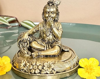 May include: A gold-coloured statue of a seated figure, with detailed ornamentation, including a crown and jewellery. The statue is on a floral-decorated base. Two yellow flowers are on either side.