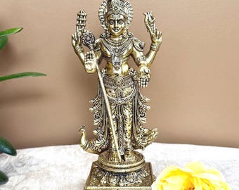 May include: A metallic gold-toned statue of a deity standing on a square base. The figure has four arms, holds a spear-like object, and is adorned with intricate jewelry and clothing. A small peacock is positioned at the feet of the figure.