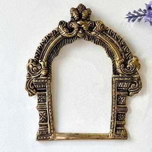 May include: An ornate, gold-coloured arch-shaped decorative frame. The frame has intricate carvings and a detailed design, with a rectangular base and a curved top. The frame is likely made of metal and is suitable for home decor.