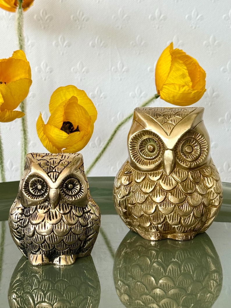 May include: Two gold-colored brass owl figurines. The larger owl is on the right and the smaller owl is on the left. The owls have detailed feather patterns and large, round eyes.