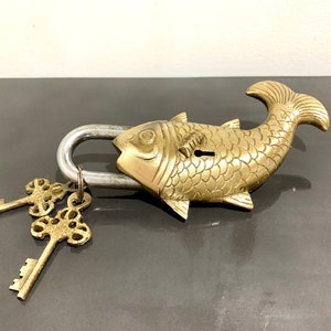 Op de afbeelding: Een messing hangslot in de vorm van een vis met twee sleutels. Het hangslot heeft een zilveren metalen beugel en de vis heeft een gedetailleerd ontwerp.