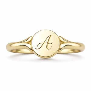 Puede incluir: Un anillo de sello dorado con una cara circular. La cara del anillo presenta la letra cursiva "A". El anillo tiene un acabado pulido y un diseño clásico. El anillo es una joya.