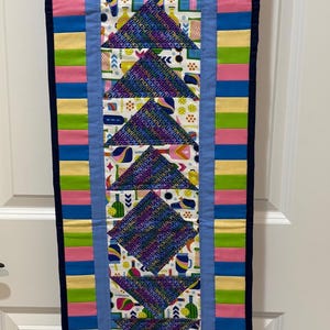 May include: A vertical quilted wall hanging featuring a central column of alternating triangles and squares with a pickleball theme. The fabric displays pickleball paddles, balls, and the word PICKLEBALL in various colors. It is bordered by colorful striped fabric in pink, blue, yellow, and green, finished with a dark navy blue binding.