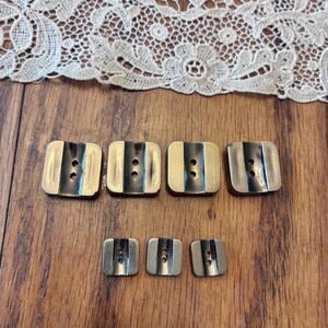 May include: A collection of rectangular buttons in various sizes. The buttons have a gold-colored metal finish with a black stripe and two holes. The buttons are arranged on a wooden surface, with a lace fabric in the background.