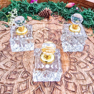 Puede incluir: Tres frascos de perfume de vidrio transparente con tapas doradas y tapones esféricos. Cada frasco cuadrado presenta un diseño impreso en blanco del ciclo de las fases lunares en la parte frontal, dispuesto en una fila.