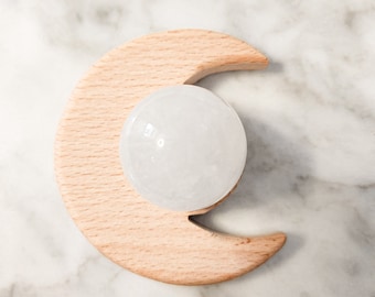 Puede incluir: Un soporte de madera clara en forma de media luna que sostiene una piedra esférica blanca, lisa y translúcida. La base presenta una textura de veta de madera visible. El texto "The Divine Moon.com" está impreso en cursiva.