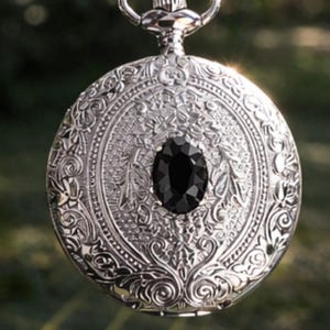 May include: Silver pocket watch with intricate floral and scrollwork design. The watch features a large, oval black gemstone in the center. The watch is suspended from a silver chain, and the background is blurred.