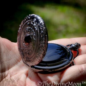 May include: Open black pocket watch with ornate detailing on the cover. The watch is held in a hand, with the open cover revealing the watch face. The watch has a black finish and a small loop for a chain.