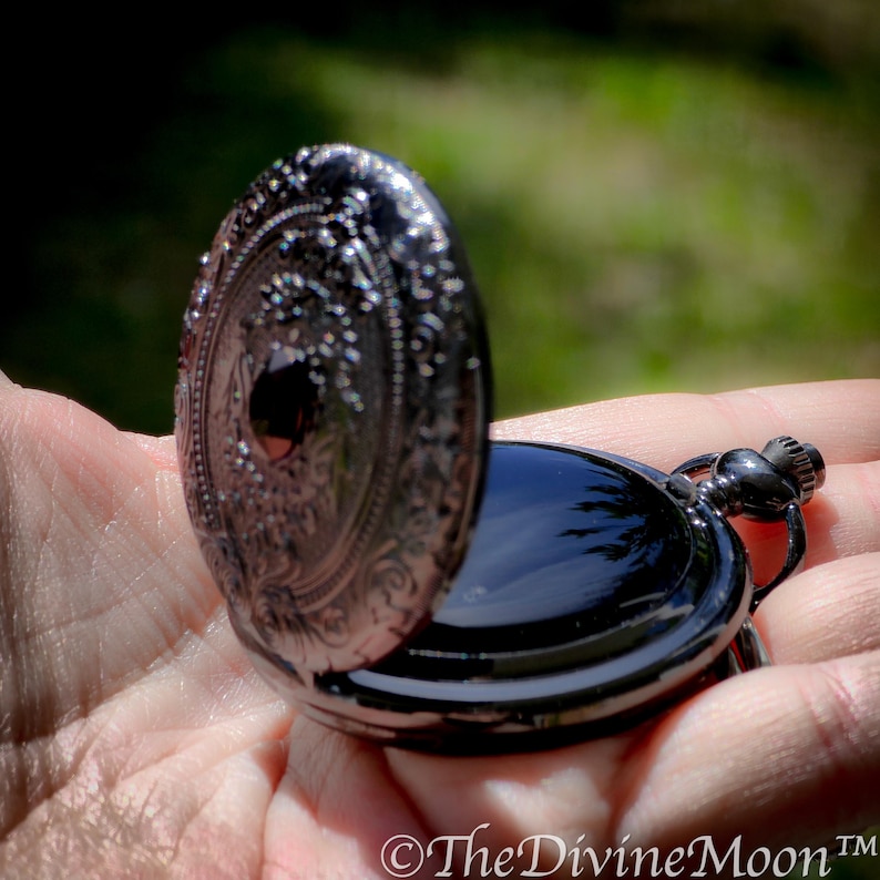 May include: Open black pocket watch with ornate detailing on the cover. The watch is held in a hand, with the open cover revealing the watch face. The watch has a black finish and a small loop for a chain.