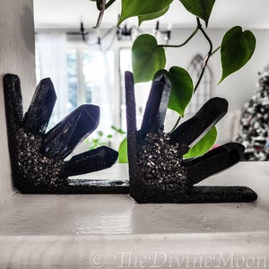 May include: Two black crystal bookends shaped like a hand with multiple crystal points. The bookends are on a white surface.