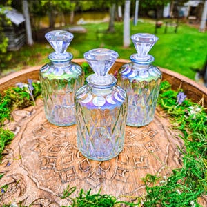 Puede incluir: Tres botellas de vidrio transparente con patrones de diamante y tapones facetados. Cada botella presenta una fila de calcoman&iacute;as blancas de las fases lunares alrededor del cuello y un brillo iridiscente en el vidrio.