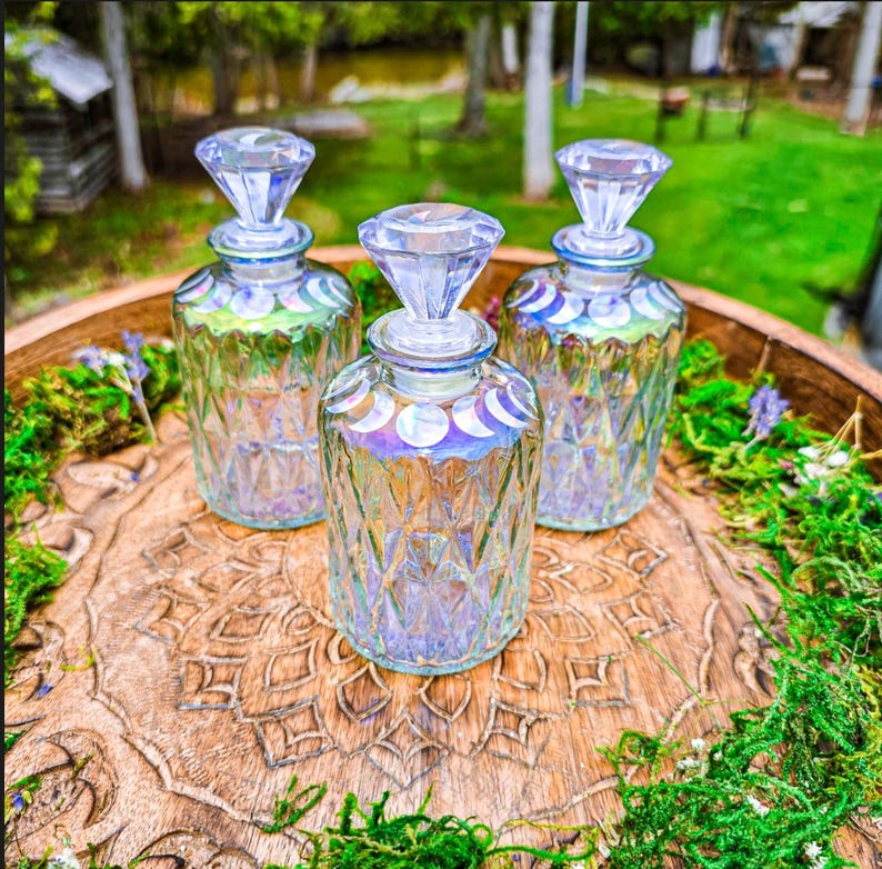Puede incluir: Tres botellas de vidrio transparente con patrones de diamante y tapones facetados. Cada botella presenta una fila de calcoman&iacute;as blancas de las fases lunares alrededor del cuello y un brillo iridiscente en el vidrio.