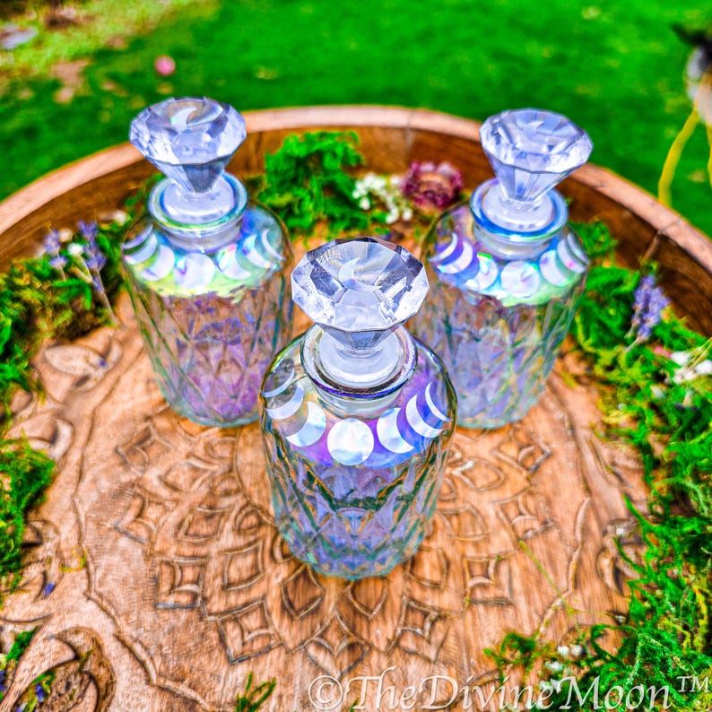 Puede incluir: Tres botellas de vidrio transparente con patrones de corte de diamante y tapones facetados. Cada botella presenta una banda de calcoman&iacute;as iridiscentes de fases lunares alrededor del cuello. El texto The Divine Moon es visible.