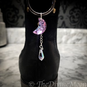 May include: A purple crystal crescent moon charm with a teardrop crystal dangling from a silver chain. The charm is attached to a black boot.