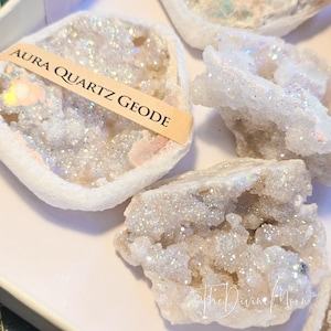 May include: Several aura quartz geode specimens are displayed, showcasing a sparkling, crystalline structure. One geode has a label that reads "Aura Quartz Geode." The geodes are white and iridescent, with a glittery appearance. The overall theme is crystals and minerals.