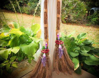 May include: Two brown twig brooms with purple and pink flower accents and crystals. The brooms are leaning against a white surface.