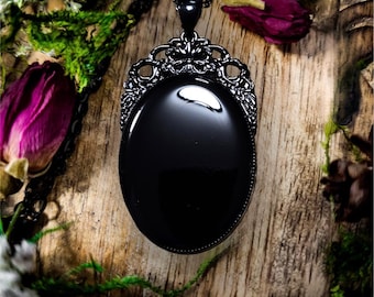 May include: Black oval pendant necklace with a delicate, ornate, black metal frame. The pendant is on a black chain and is resting on a wooden surface.