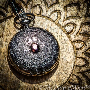 May include: A detailed, antique-style pocket watch with a dark metallic finish. The watch features an ornate, engraved design with a central, oval-shaped, dark red gemstone. The watch is suspended by a chain and rests on a wooden surface.